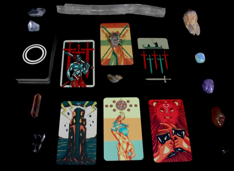 May include: Tarot card spread with ten cards laid out on a black surface. The cards are from a deck with colorful illustrations and include the Tower, the Devil, the Three of Swords, the Two of Swords, the Five of Swords, the King of Swords, the World, and the Strength card. There are also several crystals and a long, white, rectangular object on the surface.