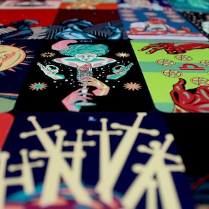 May include: A close-up of a tarot card deck with various cards spread out. The cards feature colorful illustrations and intricate designs. The cards are arranged in a grid pattern, with the top row showing the cards "VIII", "PAGE", "LIBRA", and "THE WORLD".