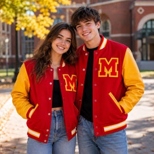Puede incluir: Una chaqueta universitaria roja con mangas amarillas, que presenta un parche con la letra M amarilla en el pecho, botones a presión blancos y ribetes acanalados a rayas en los puños y el dobladillo, sobre una camiseta negra y vaqueros.