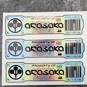 Pode incluir: Três autocolantes holográficos retangulares com o texto Property of Arasaka, um logótipo de árvore circular, um padrão de grelha e um código de barras com o texto Corporate Asset ID e ARSK-77X9-0421 impresso a preto numa superfície refletora.