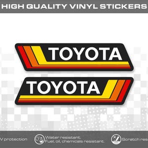 Könnte beinhalten: Zwei rechteckige Vinylaufkleber mit dem Wort TOYOTA in weißen Großbuchstaben auf schwarzem Hintergrund. Jeder Aufkleber enthält ein dekoratives Streifenmuster in Gelb, Orange und Rot. Der Text HIGH QUALITY VINYL STICKERS befindet sich oben.