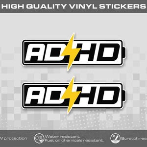 Könnte beinhalten: Zwei rechteckige Vinyl-Aufkleber mit dem Text ADHD in einer fetten schwarz-weißen Schriftart, die wie ein Batteriesymbol gestaltet ist. Ein leuchtend gelber Blitz befindet sich in der Mitte jedes Aufklebers und ersetzt den Buchstaben H.