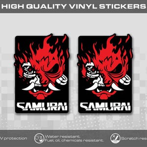 Könnte beinhalten: Zwei rechteckige Vinylaufkleber mit einer stilisierten rot-weißen Samurai-Maskengrafik auf schwarzem Hintergrund. Das Wort SAMURAI ist in weißen Großbuchstaben am unteren Rand jedes Aufklebers gedruckt, ergänzt durch flammenartige Details.