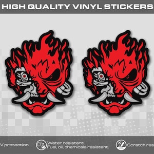 Peut inclure: Deux autocollants en vinyle identiques représentant un graphique stylisé rouge et noir d'un masque de démon mécanique avec des crocs acérés et des flammes. Le texte HIGH QUALITY VINYL STICKERS figure en haut, avec des icônes de résistance.