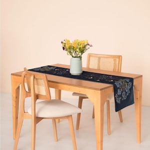 May include: A light wood dining table with two chairs featuring woven cane backrests and off-white cushions. A dark blue table runner with a floral pattern lies across the center, topped with a light green vase holding yellow flowers.