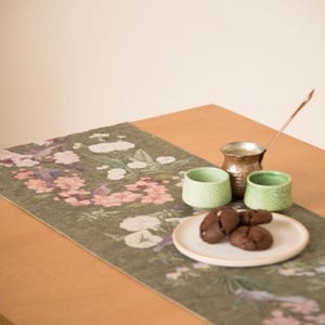 May include: A rectangular olive green table runner featuring a printed pattern of pink and white flowers and birds. On the runner sits a metallic coffee pot with a long spoon, two small light green textured cups, and a white plate with cookies.