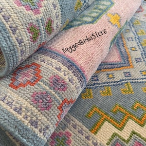 May include: A hand-tufted wool rug featuring a light blue base with geometric patterns in pink, orange, yellow, and green. The rug is partially rolled, revealing a textured pile and the text RuggedIndiaStore printed on the surface in black script.