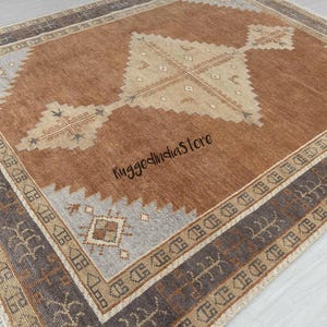 May include: A rectangular area rug featuring a large central diamond motif in beige on a brown field, surrounded by a gray border with geometric patterns and the text RuggedIndiaStore. The rug has a fringed edge and a textured surface.
