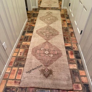 May include: A long, rectangular runner rug featuring a light beige base with repeating diamond-shaped patterns in muted brown and dusty pink tones. The rug displays the text RuggedIndiaStore printed in black near the bottom edge.
