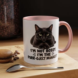 May include: A white ceramic mug with a soft pink interior and handle. The front features a graphic of a black cat with its paws crossed, accompanied by the black text: I'M NOT BOSSY. I'M THE PURR-OJECT MANAGER. The mug has a smooth, glossy finish.
