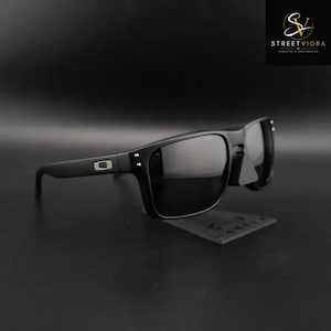 Peut inclure: Lunettes de soleil noir mat avec une monture rectangulaire et des verres teintés foncés. Les branches présentent un logo rectangulaire en argent métallique près de la charnière. La monture inclut de petits détails en clous argentés.