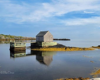 May include: A weathered wooden shed sits on a small island, connected to a pier. The shed's reflection shimmers in the calm water. The sky is a clear blue, and the scene evokes a sense of tranquility and coastal charm.