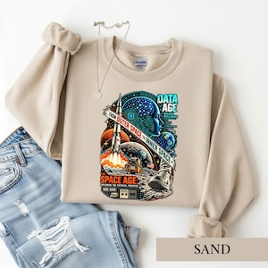 May include: A sand-colored crewneck sweatshirt featuring a graphic print of a rocket, a lunar lander, a human head silhouette with circuit patterns, and the text "DATA AGE" and "FROM OUTER SPACE TO INNER SPACE" in blue, orange, and white.