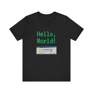 Op de afbeelding: Een zwart t-shirt met korte mouwen voorzien van de groene pixeltekst Hello, World! boven een pop-upvenstergrafiek van een computer met een geel waarschuwingspictogram en de tekst Low battery level of 2% met een selectievakje en knoppen.