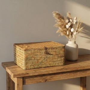 May include: A rectangular woven seagrass storage box with a hinged lid and a small wooden toggle closure. Next to the box sits a matte off-white ceramic vase containing a dried arrangement of pampas grass, lunaria, and wheat stalks.