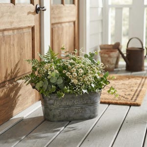 May include: An oval galvanized metal planter with the embossed text Heartful Garden Since 1972. The container is filled with a lush arrangement of green ferns, eucalyptus leaves, ivy, and small white flowers, creating a dense botanical display.