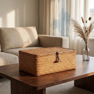 May include: A rectangular woven storage box made of light brown natural fibers. The box features a flat lid with a secure closure consisting of a dark cord loop and a small wooden toggle button centered on the front panel of the container.