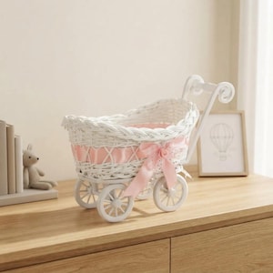 May include: A white wicker doll carriage featuring a woven basket design with a light pink satin ribbon woven through the rim. The carriage is accented with a large pink bow and a small fabric flower on the side, supported by four small white wheels.