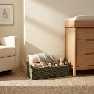 May include: A rectangular woven basket in a muted sage green color with dark brown wooden handles. The basket is filled with folded cream and light blue blankets, a small wooden bear-shaped toy, and a stackable wooden ring toy for children.