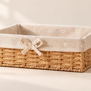 May include: A rectangular woven basket with a natural tan finish, featuring a removable beige fabric liner decorated with a small white floral pattern and a centered fabric bow detail on the front edge of the basket rim.