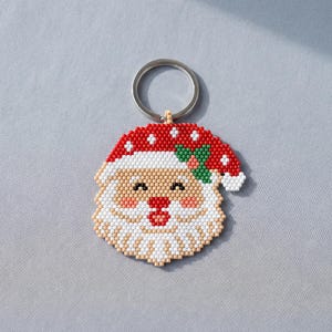 Puede incluir: Un llavero de cuentas con un diseño de cara de Papá Noel. El Santa tiene un gorro rojo con lunares blancos y un detalle de hoja de acebo verde, cara color melocotón, mejillas sonrosadas, nariz roja y una barba blanca de cuentas cuadradas.