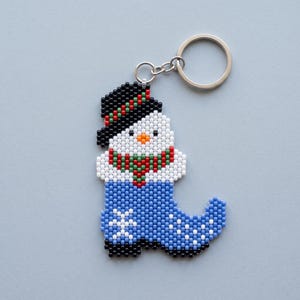 May include: A beaded keyring featuring a snowman wearing a black top hat with a red and green band, a red and green scarf, and a blue boot-shaped base decorated with a white snowflake pattern, attached to a silver-toned metal split ring.