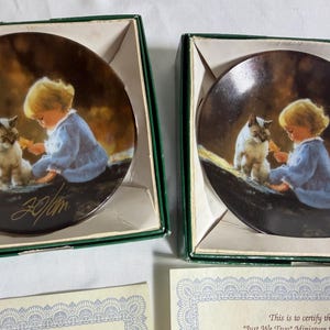 May include: Two circular decorative plates titled Just We Two, each featuring a blonde child in a blue dress interacting with a Siamese cat. The plates are housed in individual green boxes, with one plate displaying a gold signature on the surface.