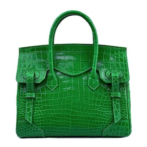Puede incluir: Un bolso de mano de color verde vibrante con una textura brillante de piel de cocodrilo. El bolso incluye dos asas superiores, un cierre de solapa y dos correas decorativas verticales en el panel frontal. El diseño es estructurado y rectangular.
