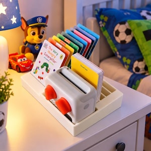 Puede incluir: Un reproductor multimedia de plástico blanco con dos perillas naranjas y una ranura para insertar tarjetas. El dispositivo está colocado en una base rectangular que sostiene una colección de tarjetas de historias coloridas, incluida una titulada The Very Hungry Caterpillar.