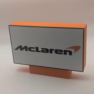 May include: A rectangular sign with a bright orange frame and a white face featuring the black McLaren logo and an orange swoosh graphic. The sign is mounted on a matching orange rectangular base, creating a stable, upright display piece.