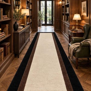 Peut inclure: Un tapis de couloir rectangulaire doté d'un large panneau central de couleur crème, d'une bordure marron moyen et d'un contour extérieur noir uni. Le tapis est placé dans le sens de la longueur sur un parquet, s'étendant vers une porte au fond.