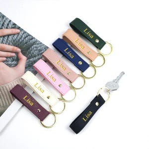 May include: A collection of seven rectangular faux leather keychains in dark green, beige, navy blue, dusty pink, light pink, cream, and dark purple. Each keychain features the name Lisa in gold lettering, a gold metal rivet, and a gold ring.