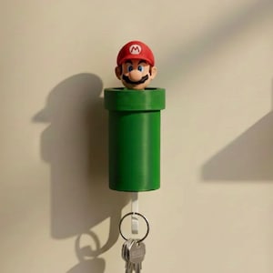 Puede incluir: Un soporte de pared para llaves que presenta una tubería cilíndrica verde con una figura de Mario emergiendo de la parte superior. La tubería está unida a un gancho blanco que sostiene un llavero de metal plateado con varias llaves.