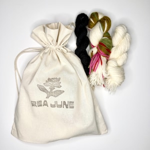 May include: A cream-colored cotton drawstring bag with the text REA JUNE and a flower graphic printed in gray. Next to the bag are three skeins of yarn: one solid black, one variegated with white, green, pink, and red, and one solid off-white.
