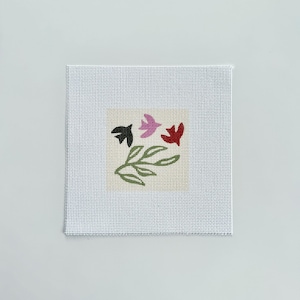 May include: A square needlepoint canvas featuring a central design of three small birds in black, pink, and red, positioned above a green outlined leafy branch. The canvas has a white grid pattern and a plain white border surrounding the design.