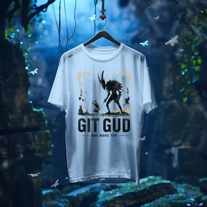 Può includere: Una t-shirt bianca a maniche corte con una stampa grafica che raffigura un piccolo personaggio di fronte a una figura alta e cornuta. Il testo "GIT GUD" è stampato in grandi lettere scure sotto l'illustrazione, con "ONE MORE TRY" sotto.