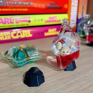 May include: A clear, decorative bottle stopper with a cartoon character design, filled with red liquid, sits on a black base. A green, star-shaped object and a black geometric shape are also visible. Books with colorful spines are in the background.