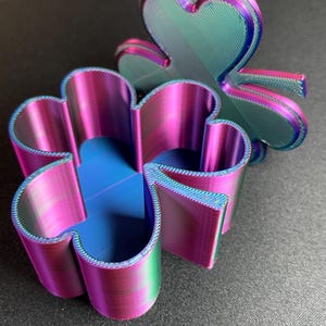 May include: A three-dimensional, iridescent, clover-shaped trinket box with a hinged lid. The box is made of plastic and has a metallic finish with shades of pink, blue, and green.