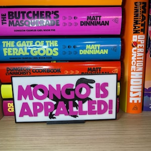Puede incluir: Un letrero rectangular blanco con el texto "MONGO IS APPALLED!" en rosa y una silueta de dinosaurio negra. El letrero est&aacute; frente a una estanter&iacute;a de libros coloridos con t&iacute;tulos como "The Eye of the Bedlam Bride" y "The Butcher's Masquerade".
