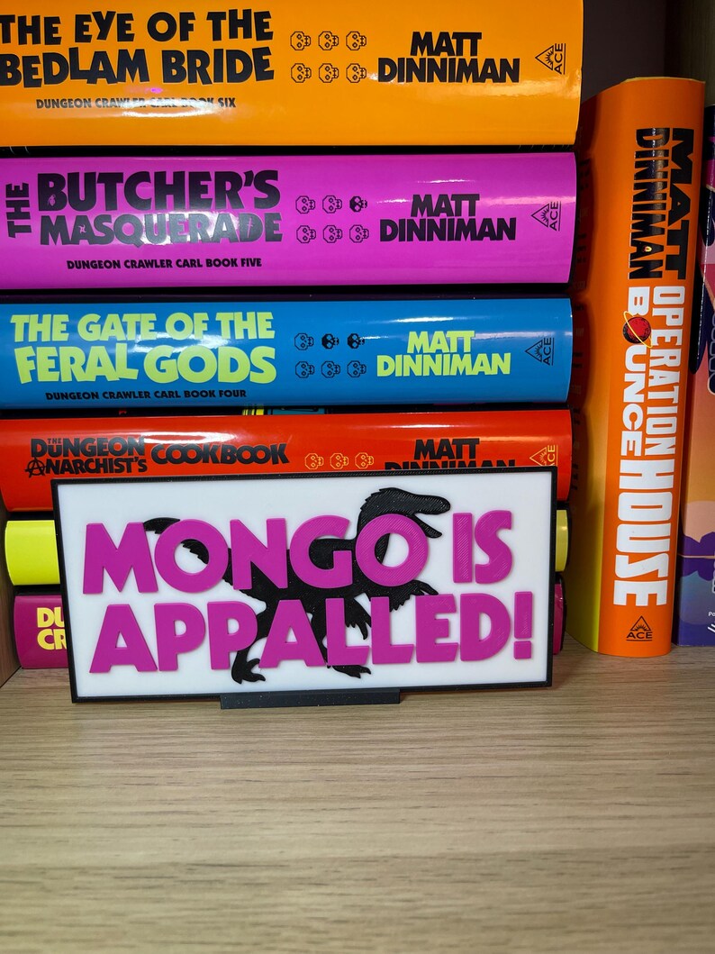 Puede incluir: Un letrero rectangular blanco con el texto "MONGO IS APPALLED!" en rosa y una silueta de dinosaurio negra. El letrero est&aacute; frente a una estanter&iacute;a de libros coloridos con t&iacute;tulos como "The Eye of the Bedlam Bride" y "The Butcher's Masquerade".