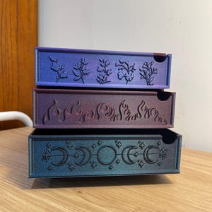 May include: Three stacked decorative boxes with intricate designs. The top box is blue with a floral pattern, the middle box is purple with a flame design, and the bottom box is teal with moons and stars.