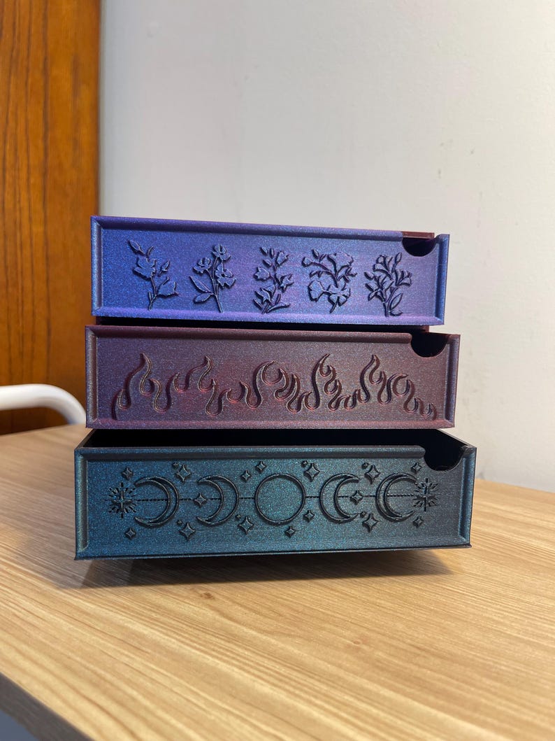 May include: Three stacked decorative boxes with intricate designs. The top box is blue with a floral pattern, the middle box is purple with a flame design, and the bottom box is teal with moons and stars.