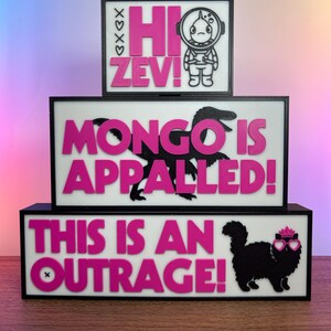 Puede incluir: Carteles de tres niveles, con marco negro, fondos blancos y texto rosa. El letrero superior dice "Hi Zev!" con un astronauta de dibujos animados. El letrero del medio dice "Mongo is Appalled!" con una silueta de dinosaurio. El letrero inferior dice "This is an Outrage!" con un gato.