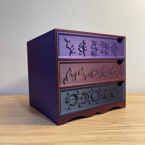 May include: A small, three-drawer storage chest in shades of purple and burgundy. Each drawer features a unique embossed design: floral, flames, and phases of the moon. The chest is made of a solid material and sits on a wooden surface.
