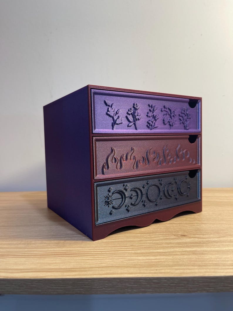 May include: A small, three-drawer storage chest in shades of purple and burgundy. Each drawer features a unique embossed design: floral, flames, and phases of the moon. The chest is made of a solid material and sits on a wooden surface.