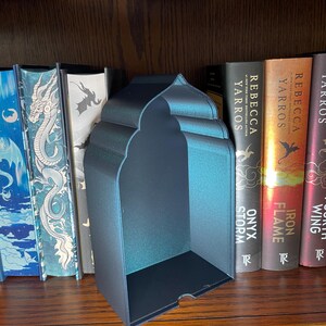 Op de afbeelding: Een zwarte metalen boekensteun met een gewelfde bovenkant en een rechthoekige basis. De boekensteun staat op een houten plank met verschillende boeken. De boekensteun heeft een lichte iriserende glans.