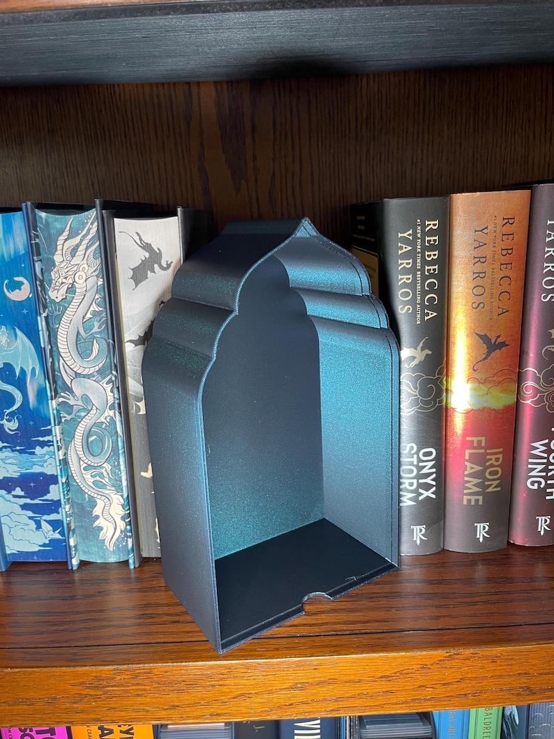 Op de afbeelding: Een zwarte metalen boekensteun met een gewelfde bovenkant en een rechthoekige basis. De boekensteun staat op een houten plank met verschillende boeken. De boekensteun heeft een lichte iriserende glans.