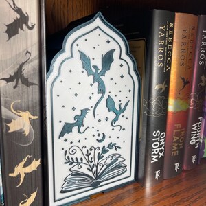 Op de afbeelding: Een witte en teal draak-thema boekensteun met draak silhouetten en een decoratief open boek ontwerp. De boekensteun staat tussen boeken op een houten plank. De boeken hebben titels als "Onyx Storm" en "Iron Flame".