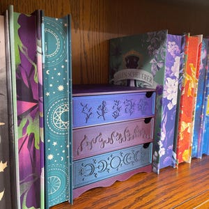 May include: A wooden bookshelf with a collection of books and a decorative three-drawer storage unit. The drawers are purple and teal with etched botanical and celestial designs. The book spines are colorful with floral and geometric patterns.