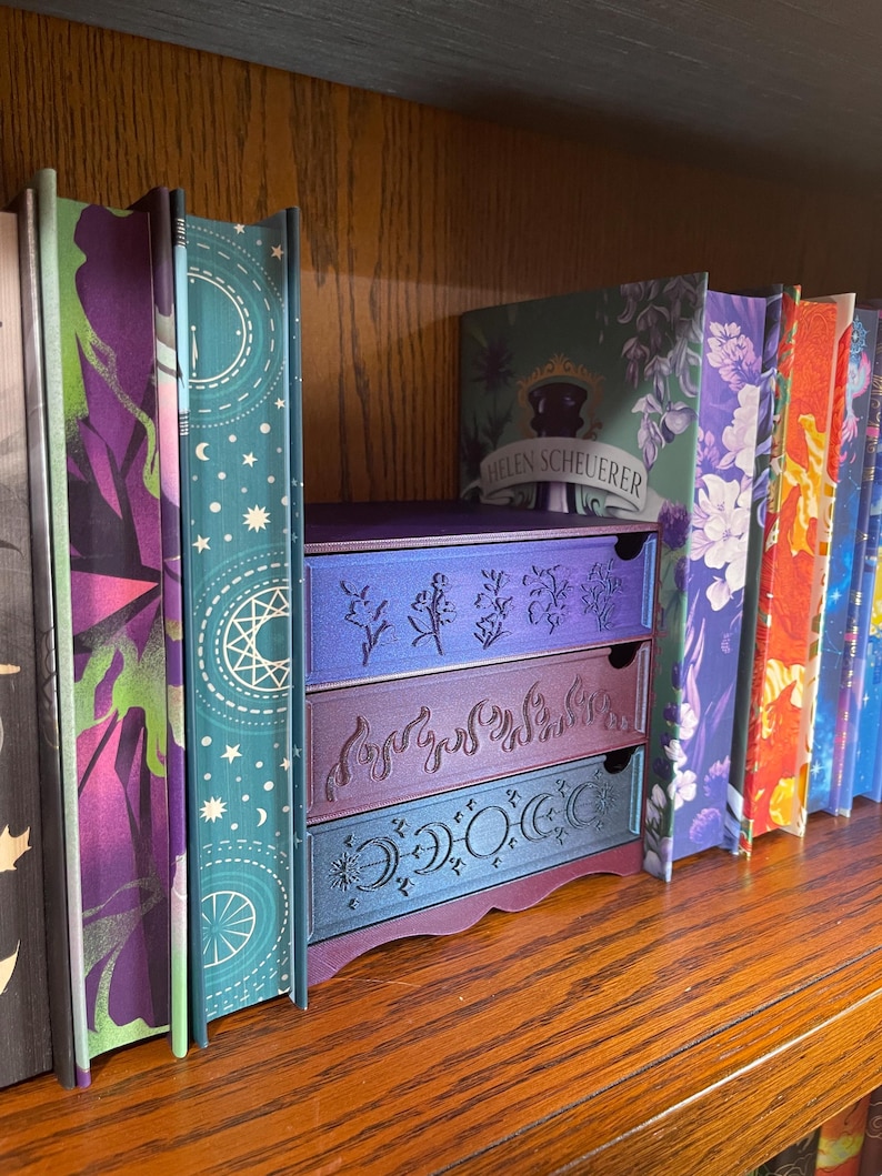 May include: A wooden bookshelf with a collection of books and a decorative three-drawer storage unit. The drawers are purple and teal with etched botanical and celestial designs. The book spines are colorful with floral and geometric patterns.